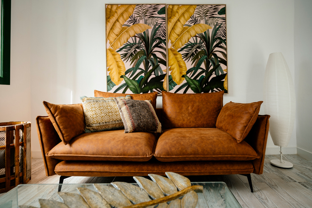 Canapé en cuir marron avec coussins décorés, tableau avec feuilles exotiques derrière.