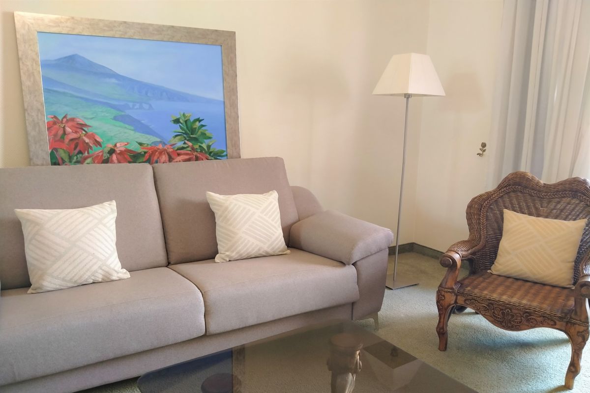 Canapé beige avec coussins, tableau de paysage de Tenerife et fauteuil en rotin.