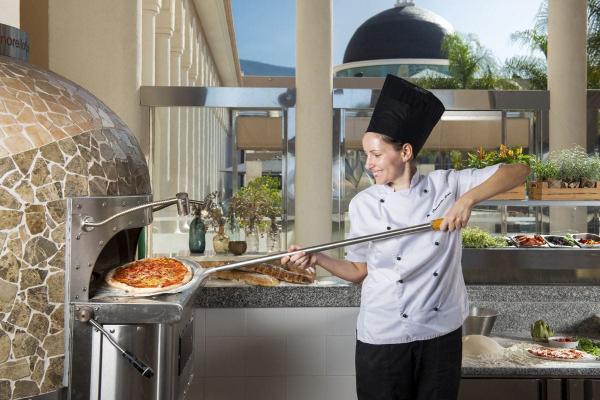Chef sortant une pizza du four à Tenerife, entourée de plantes et ingrédients frais.