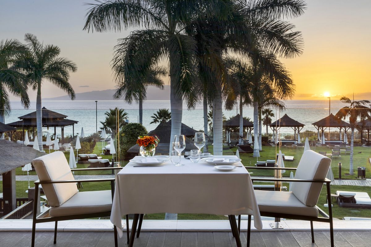 Table dressée avec vue sur le coucher de soleil à Tenerife, avec palmiers et océan en arrière-plan.