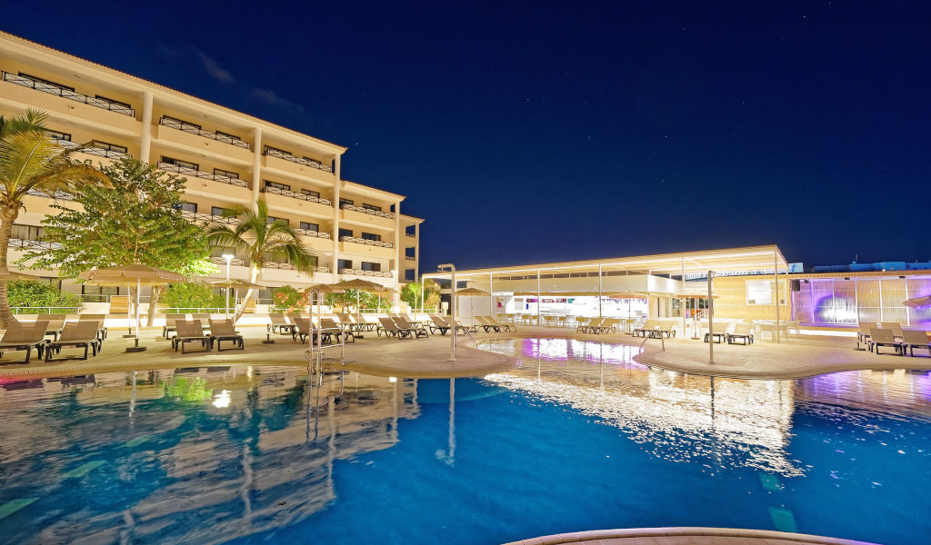 Piscine d'un hôtel éclairé la nuit à Tenerife avec palmiers et chaises longues.