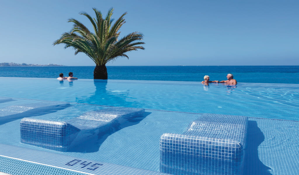 Piscine à débordement avec vue sur la mer à Tenerife, palmier et deux nageurs visibles.