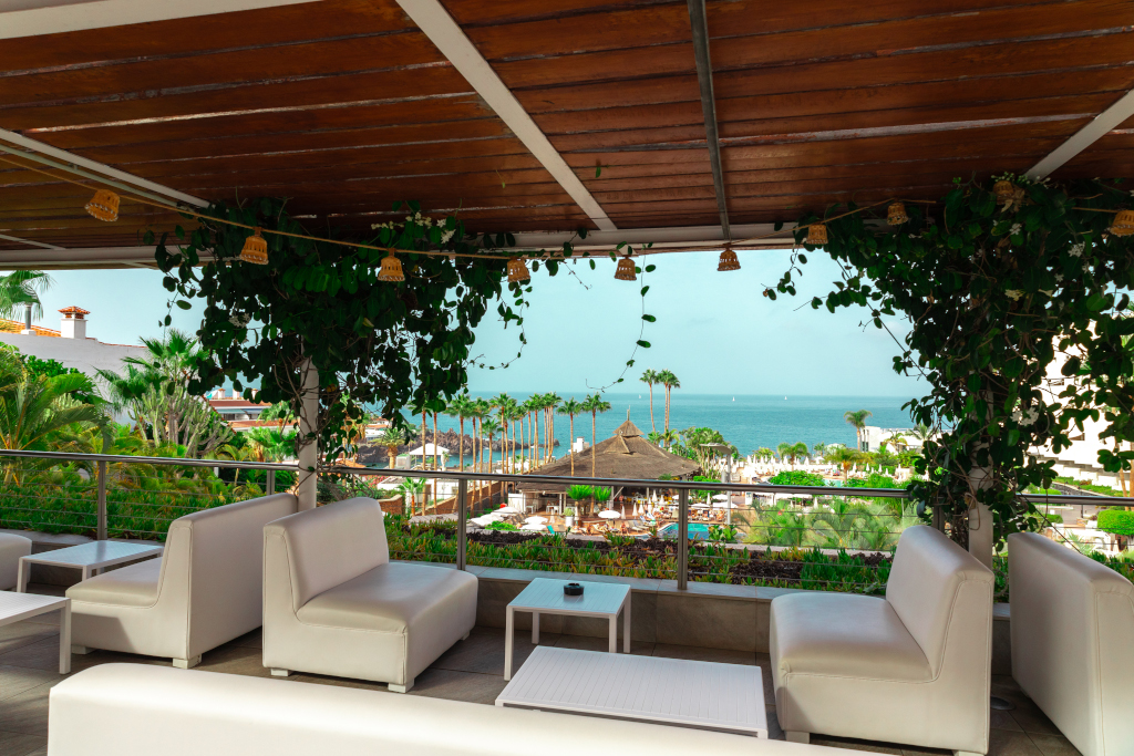 Vue sur la mer depuis une terrasse à Costa Adeje, Tenerife, avec palmiers et sièges blancs.