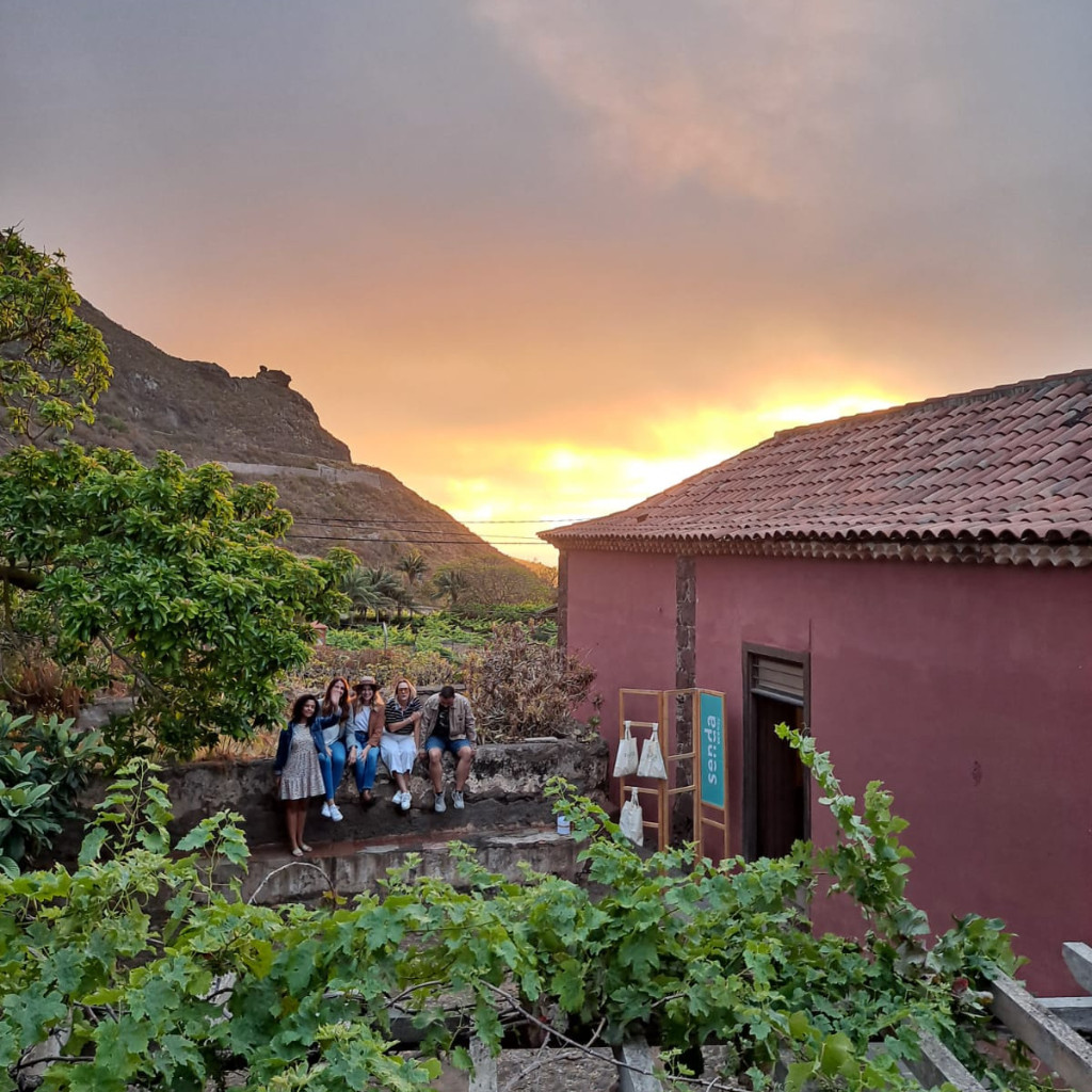 Groupe de personnes assises au coucher du soleil dans un jardin à Tenerife, avec un panneau indiquant un chemin.