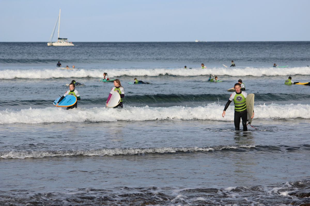 Des surfeurs avec des planches à Playa de Las Américas, Tenerife, avec un voilier en arrière-plan.