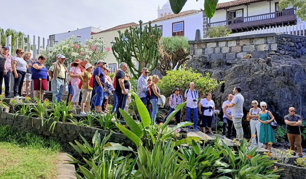 Groupe de personnes se promenant dans un jardin d’un endroit à Tenerife, entouré de plantes et d’architecture locale.