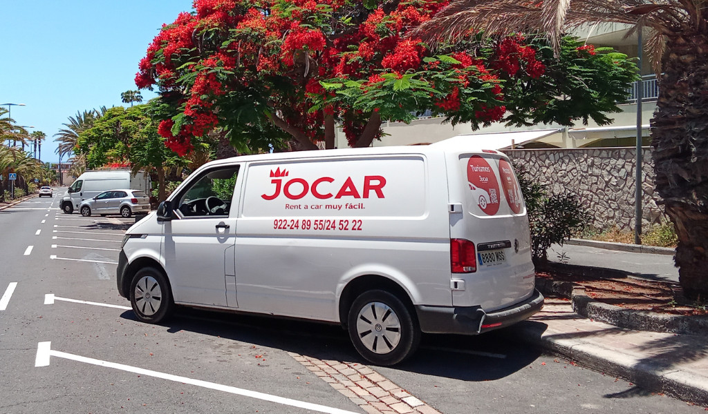 Fourgonnette de location Jocar à Santa Cruz de Tenerife garée à côté d’un arbre aux fleurs rouges.