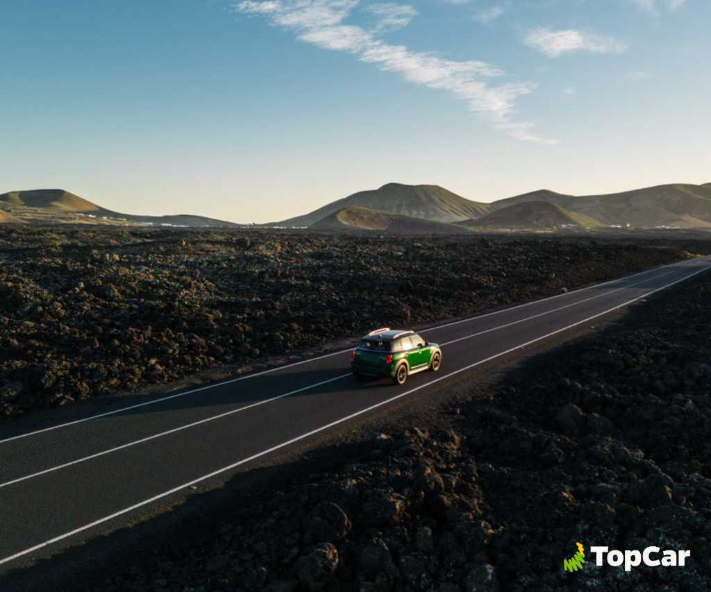 Voiture verte circulant sur la route entre des paysages volcaniques à Tenerife. Logo TopCar.