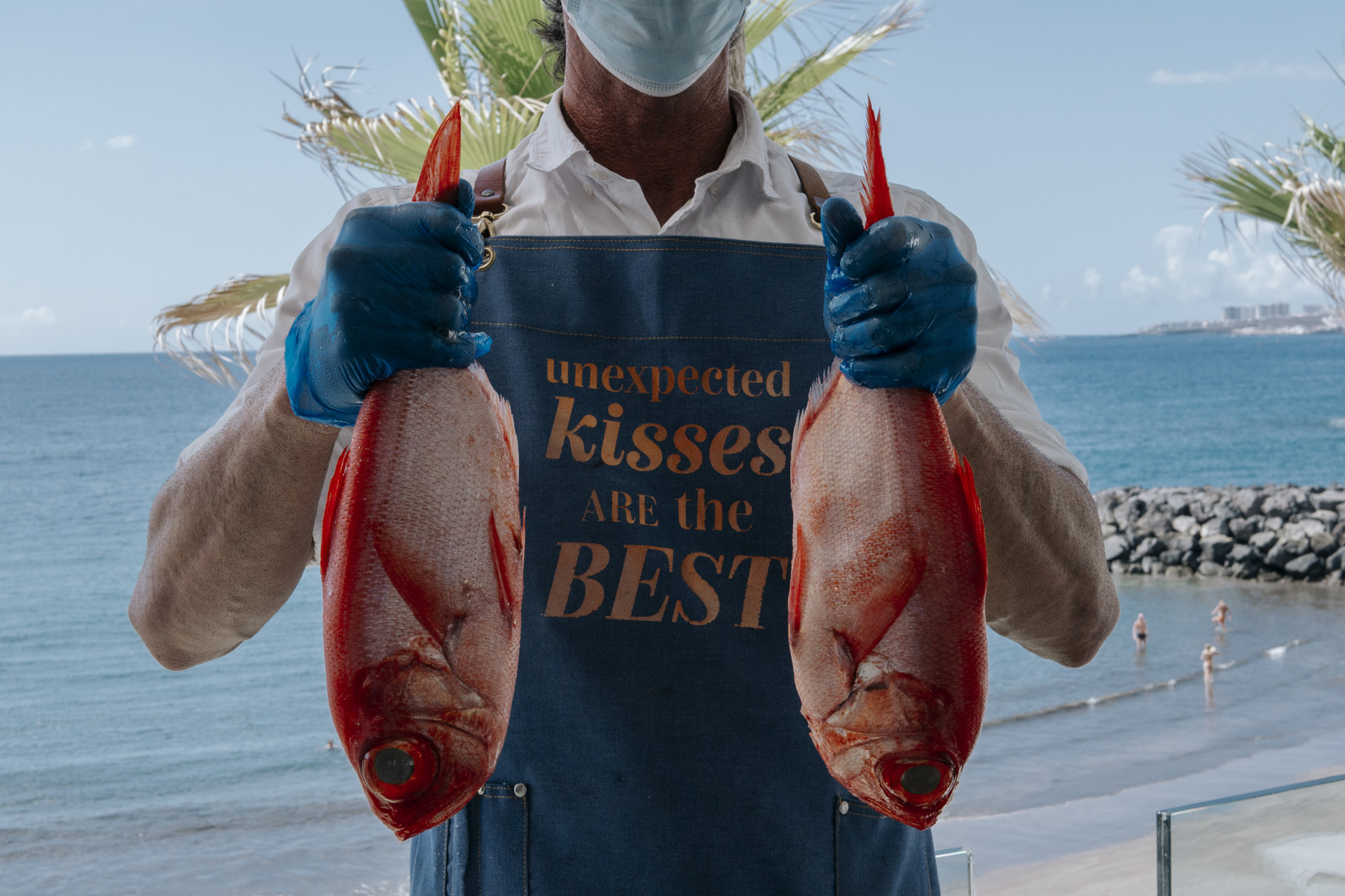 Une personne tenant deux poissons avec un tablier disant que les baisers inattendus sont les meilleurs de Tenerife.