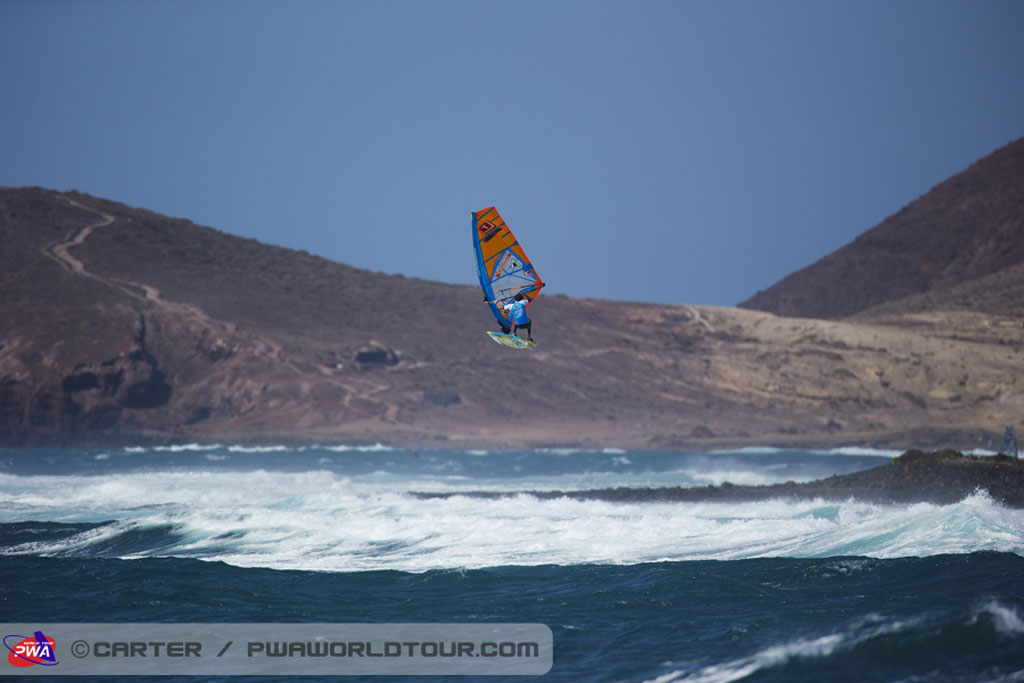 Un planchiste à voile dans les airs à El Médano, Tenerife, avec vagues et montagnes en arrière-plan.