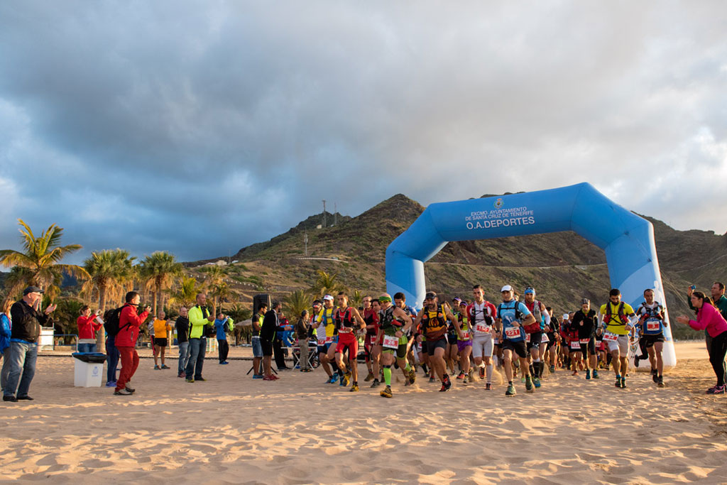 Des coureurs qui commencent un marathon sur la plage de Las Teresitas, à Tenerife, sous une arche bleue.