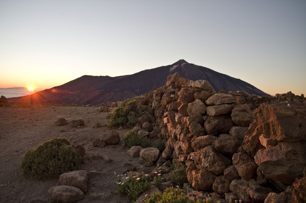 Le lever du soleil sur El Teide, à Tenerife, avec des rochers et de la végétation au premier plan.