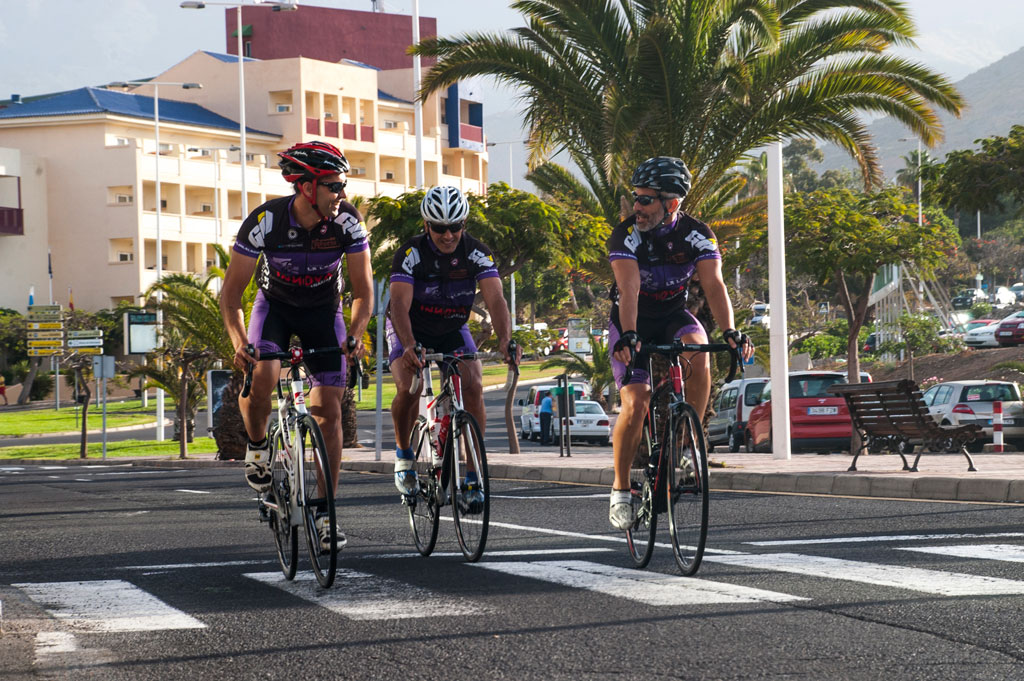 Des cyclistes traversent une route à Tenerife avec un bâtiment en arrière-plan et des palmiers autour.