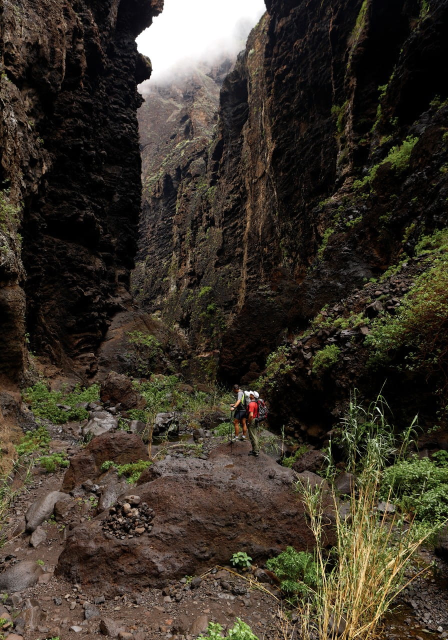 Deux personnes traversant le Barranco de Masca à Tenerife entourées de hauts murs rocheux.